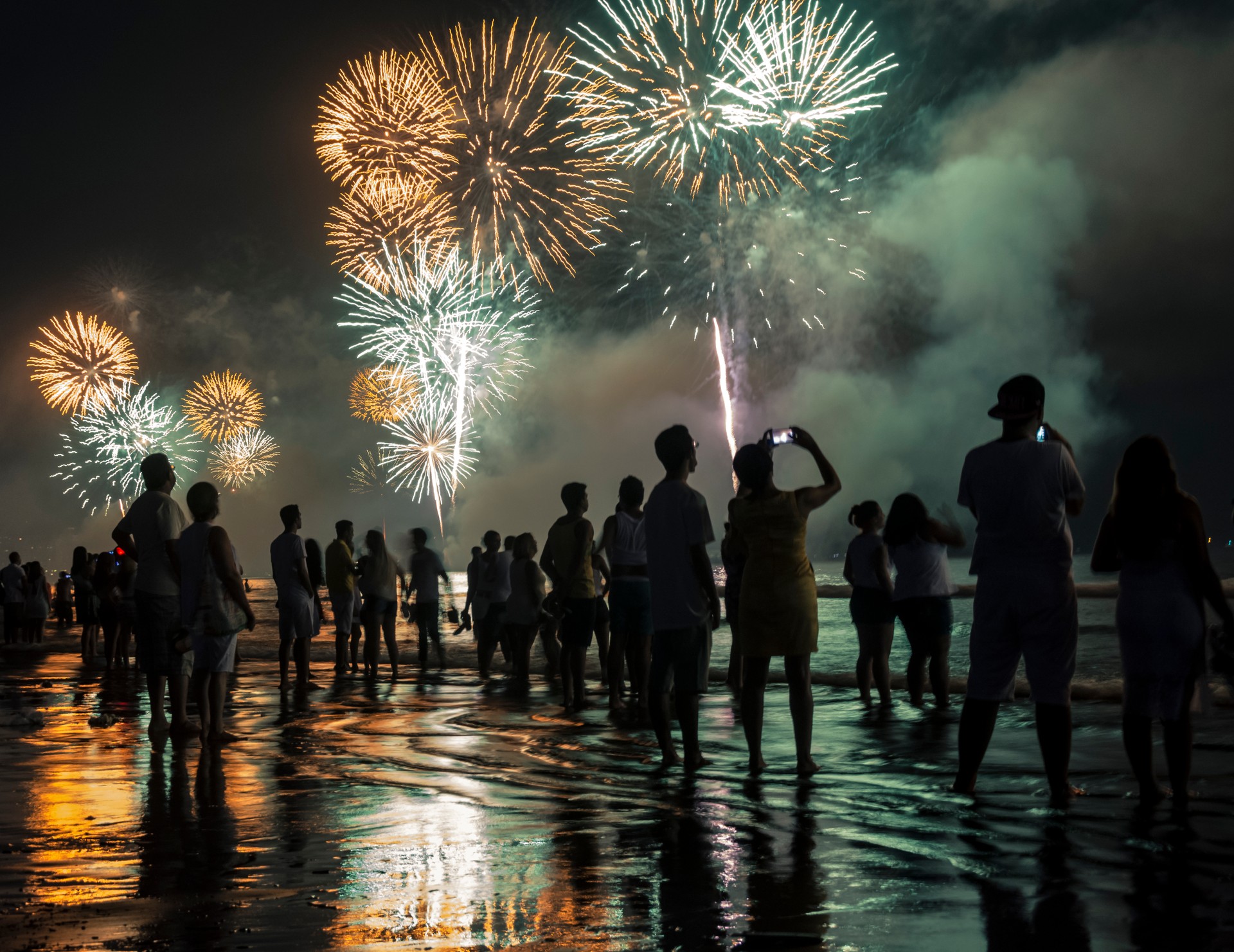 Celebração de Ano Novo na praia. Pessoas assistindo aos fogos de artifício à beira da água.